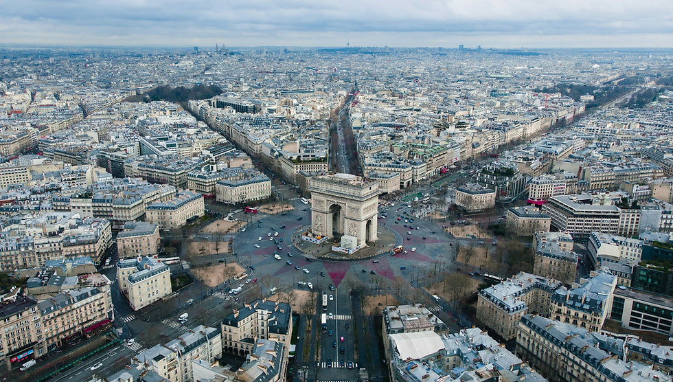 Aerial view of Paris with the Arc de Triomphe at center, surrounded by dense city streets under a cloudy sky.