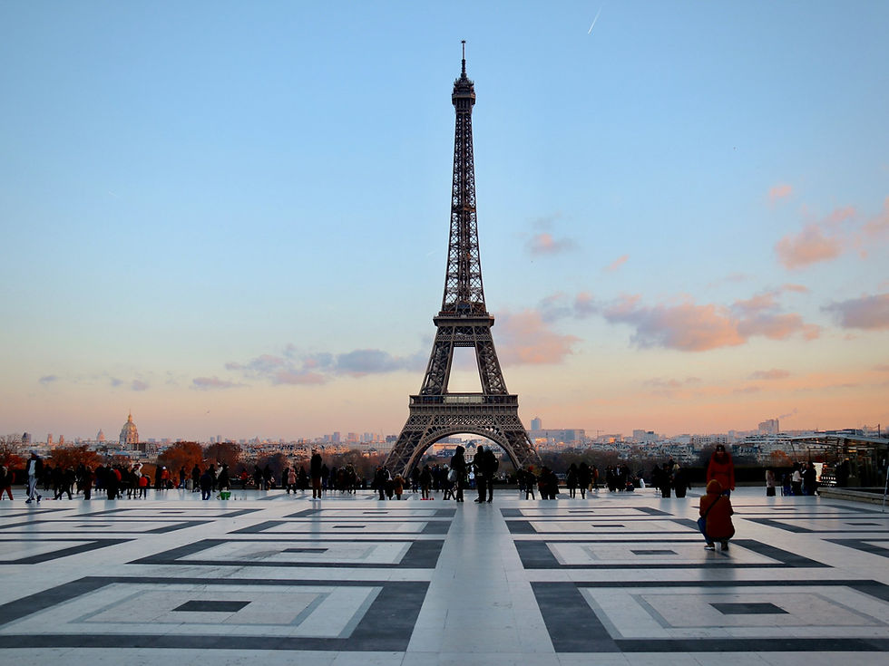 People gather on a patterned plaza before the Eiffel Tower at sunset, with pink clouds and Paris skyline in the background.