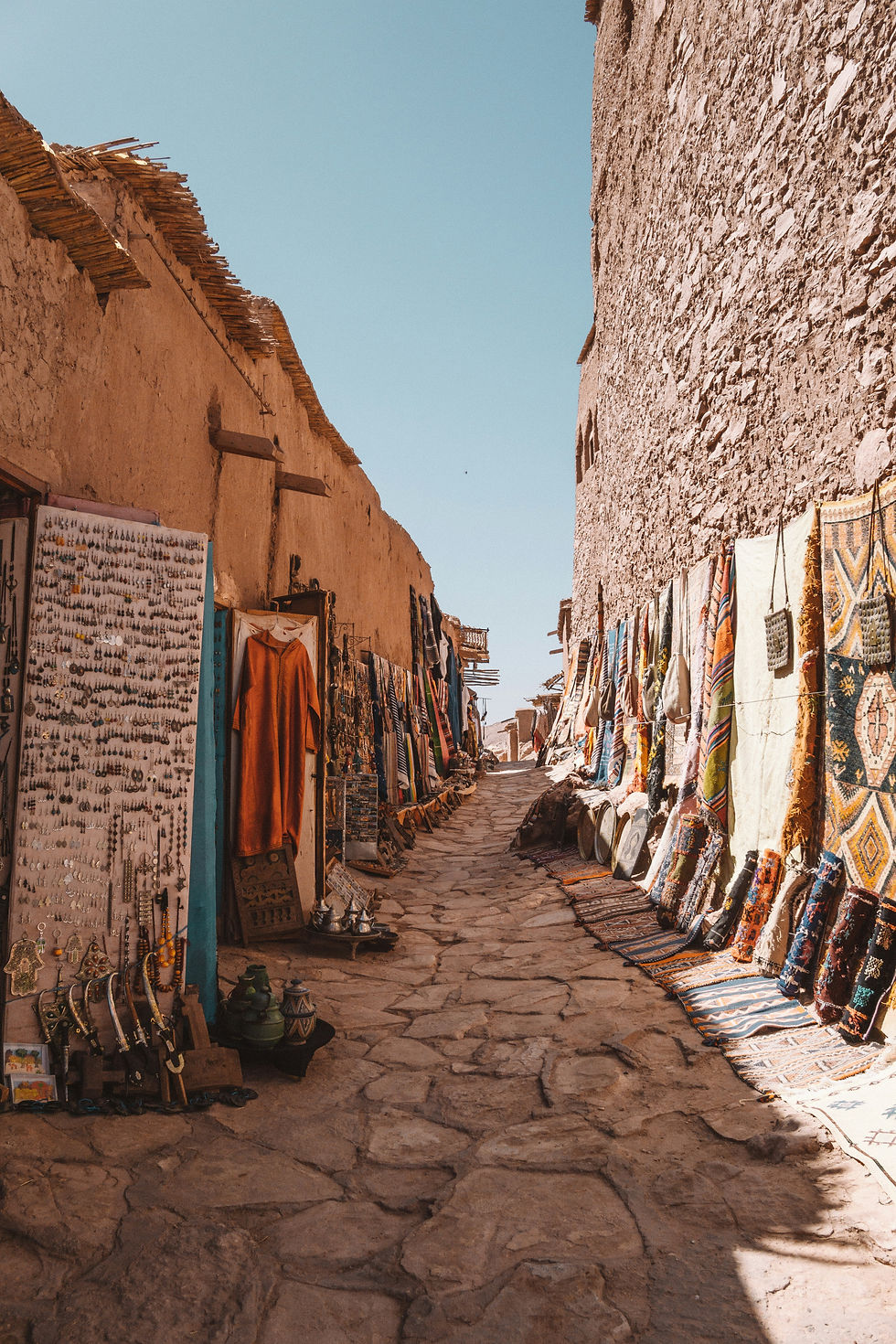 Narrow stone street with hanging colorful rugs and trinkets for sale. Rustic walls and blue sky create a vibrant, historic market vibe.