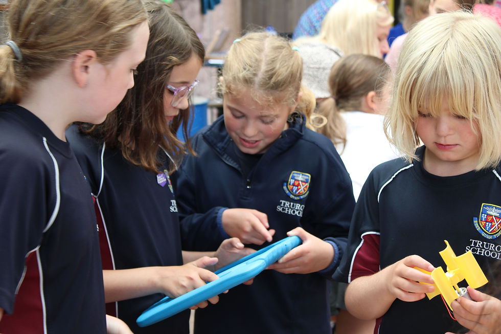 Children in navy shirts examine a blue tablet and a yellow object in a busy setting. "Truro School" logo visible, focus and curiosity evident.