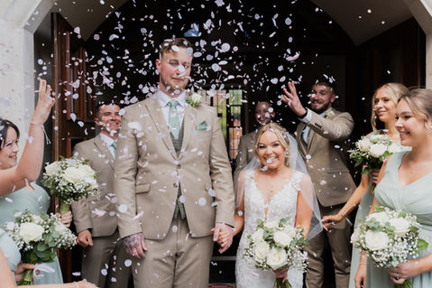 Bride and groom smiling through falling confetti outside a traditional church wedding venue.