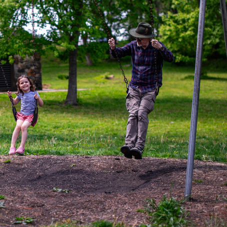 Older man wearing a hat swings alongside a laughing young girl on playground swings, both mid-air on a sunny day in the grass