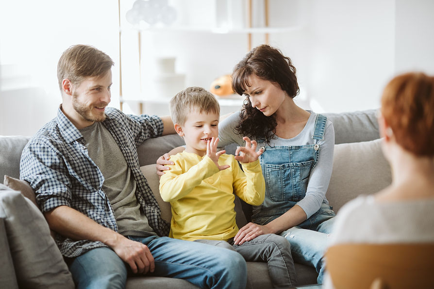 A family sitting on a sofa watching their young son gesture with his hands during a family therapy session.