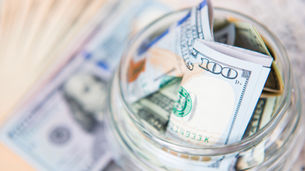 Jar filled with U.S. dollar bills, focusing on a $100 note, set on a blurred background of more cash.