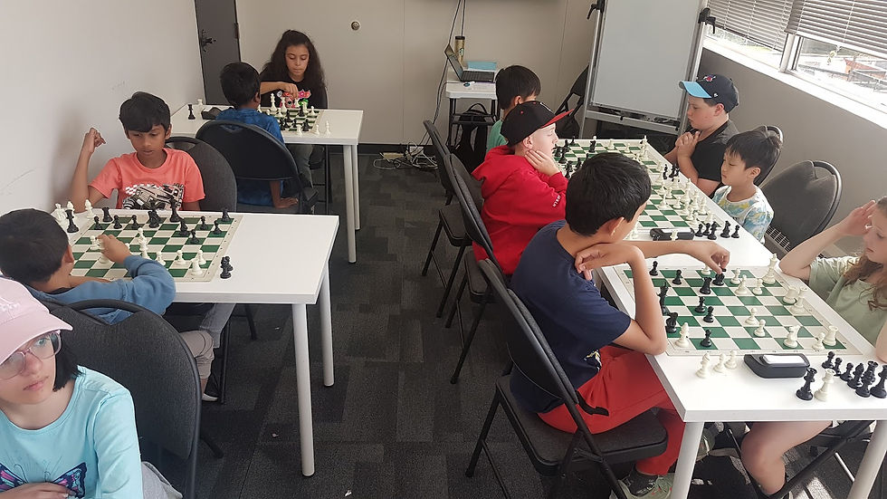 Children play chess on white tables in a room with large windows. They appear focused and engaged. One child wears a bright red hoodie.