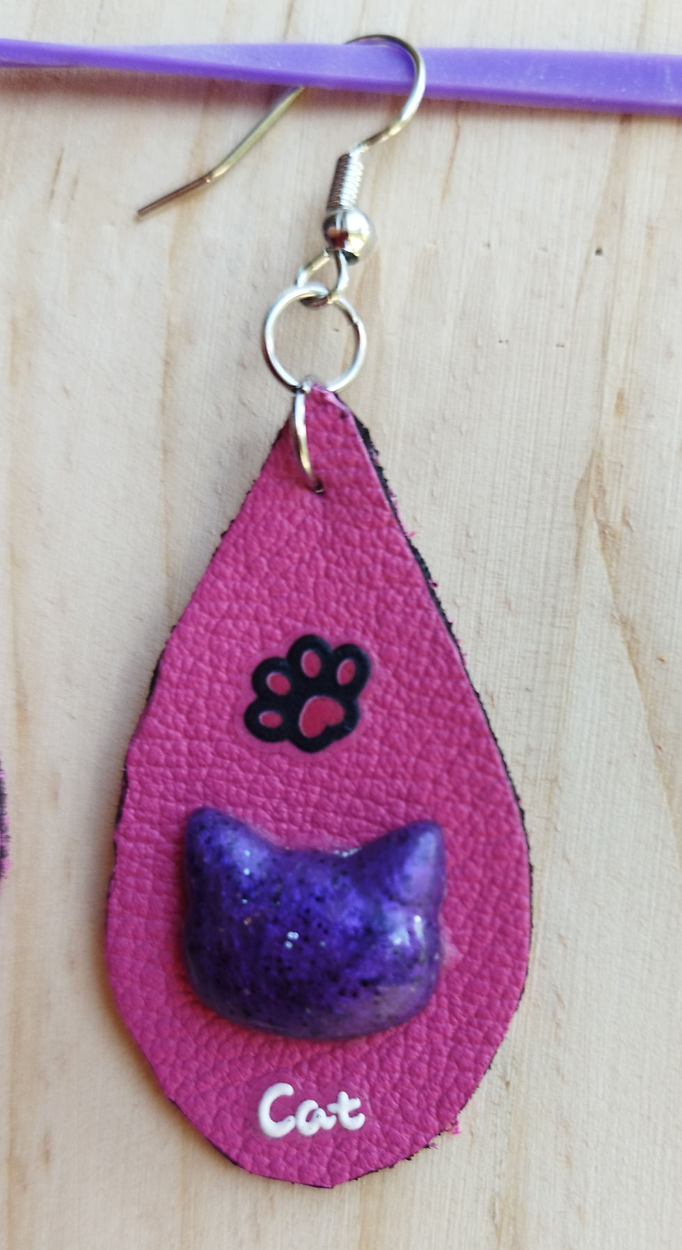 Boucles d'oreilles forme goutte cuir rose avec une tête de chat violette une petite patte noire et le mot "cat" en blanc.