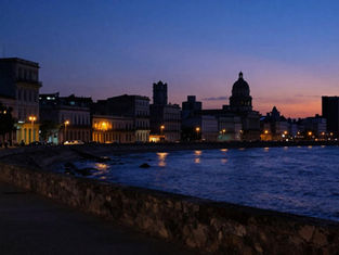 Havana's Malecón seawall and colonial skyline unusually dimmed during a power outage at blue hour, with only scattered warm lights across the city, October 2026.