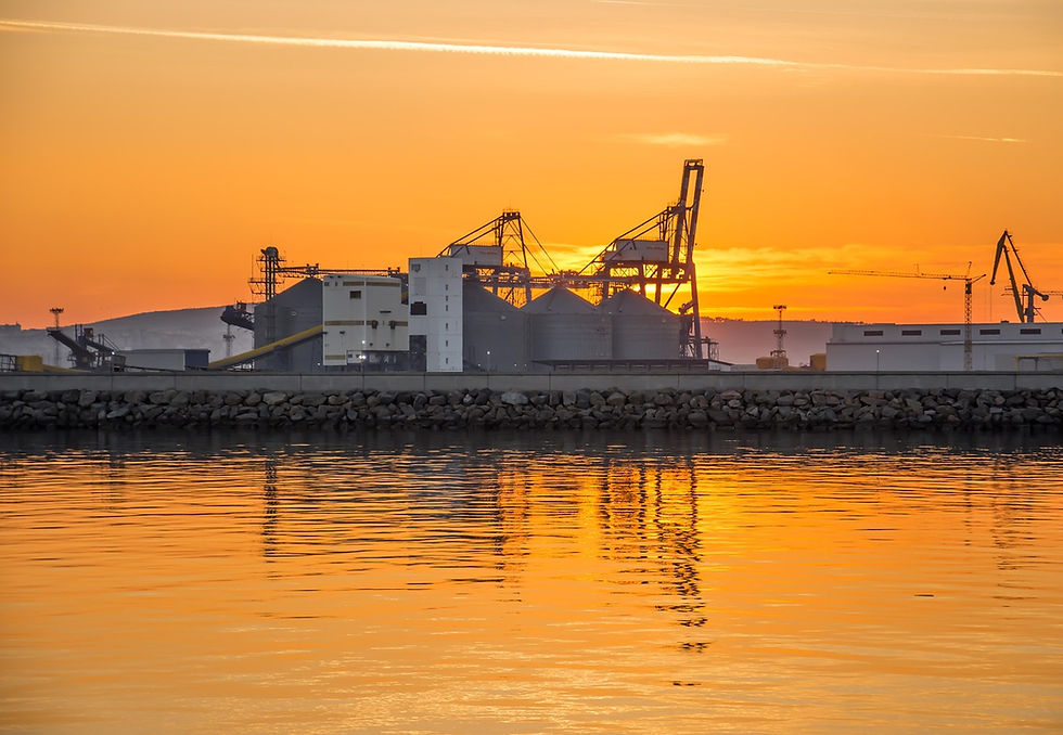 Porto industrial ao pôr do sol, com as silhuetas dos guindastes contra um céu alaranjado. As águas calmas refletem as tonalidades vibrantes, criando uma cena serena.