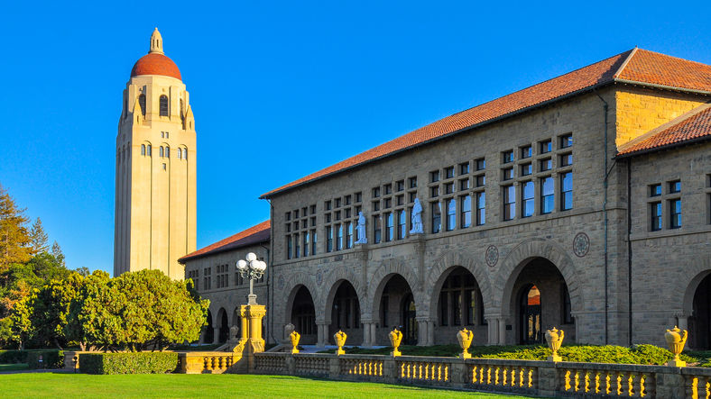 Historic university campus with a tall tower and arched building. Clear blue sky, green lawns, and bright sunlight convey a serene atmosphere.