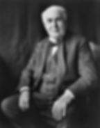 Thomas Edison in a suit sits on a chair against a dark backdrop, looking slightly serious and contemplative. Black and white photograph.