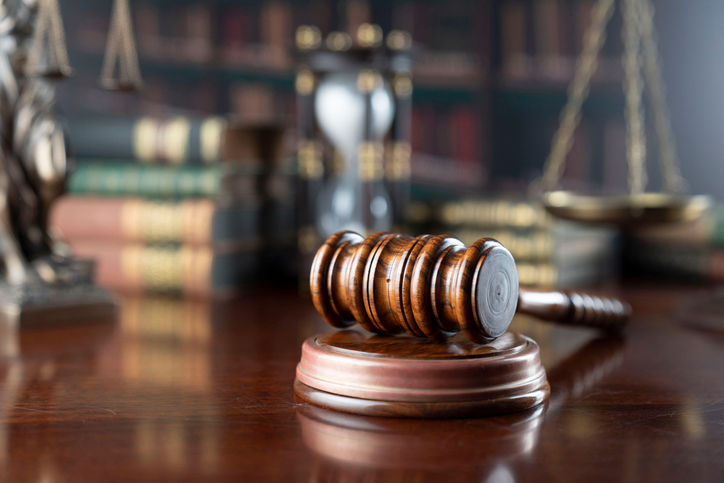 Gavel on wooden table with blurred law books, scales, and hourglass in background. Warm lighting creates a formal, serious ambiance.