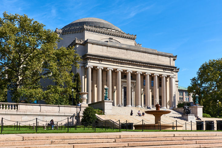 Grand classical building with columns, located in a sunny park setting. People sit on steps; text on facade reads "Library of Columbia Univ."