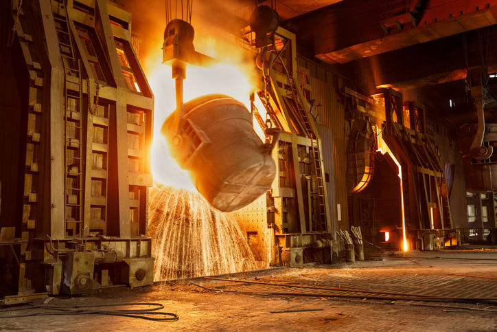 Molten metal pours from a large ladle in a vibrant, fiery steel mill. The industrial setting is illuminated with intense orange and yellow light.