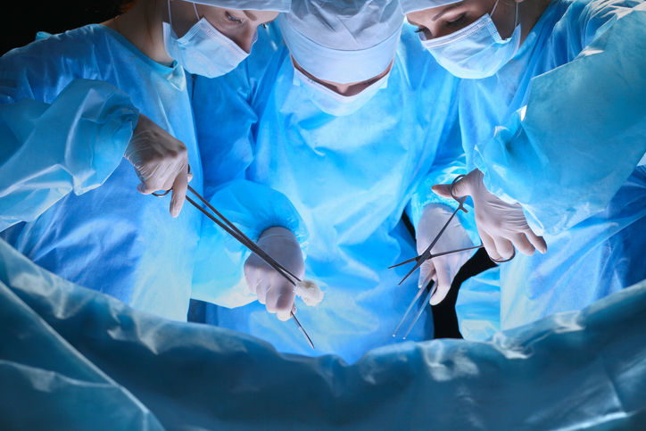 Surgeons in blue scrubs and masks perform surgery under bright lights. Three hands hold instruments, focused and collaborative atmosphere.