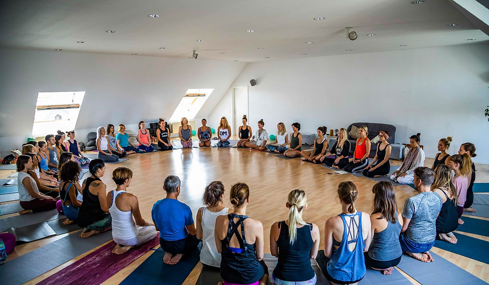 A diverse group of people sit in a circle on yoga mats in a bright room with wooden floors, engaging in a meditative practice. Mood is calm.