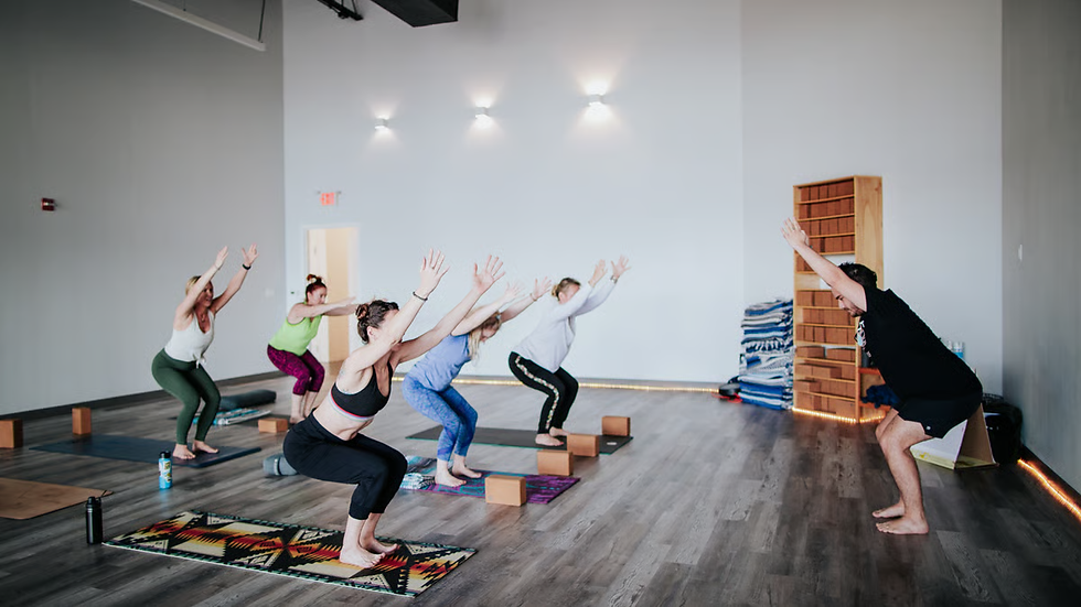 People practice yoga in a studio, holding a pose with arms raised. They are on mats with wooden floors and a minimalistic background.
