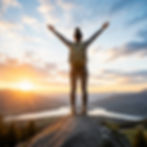 Person standing on a mountaintop at sunrise with arms raised toward the sky.