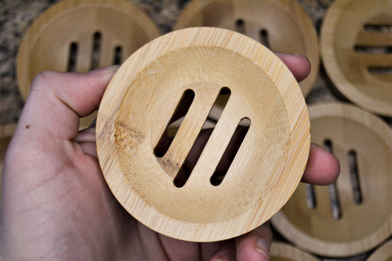 Hand holding a round, wooden soap dish with diagonal slots and other dishes in the background