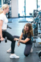 In einem modernen Trainingsbereich des LOVT Fitnessstudios korrigiert eine Personal Trainerin in schwarzem Sportoutfit die Ausführung einer Kniebeuge mit Kurzhantel bei einer blonden Frau im weißen Shirt. Die Kundin hält in der linken Hand eine Hantel, während die Trainerin mit Blick auf die Haltung das Knie leicht berührt und Anweisungen gibt. Im Hintergrund sind LOVT-Gewichte und Fitnessgeräte sichtbar. Die Szene zeigt gezieltes Krafttraining, funktionelles Coaching und persönliche Betreuung im Fitnessstudio.