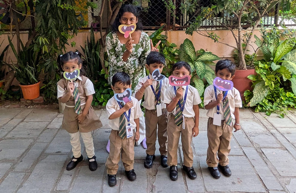 Teacher and five schoolchildren in uniforms pose outdoors, holding colorful monster-mouth cutouts amid garden greenery.