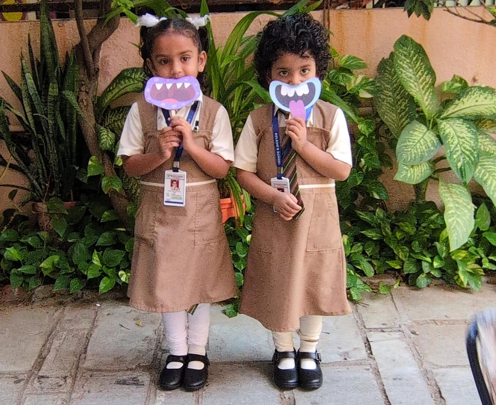 Two schoolgirls in brown uniforms hold monster-mouth cutouts in a leafy school courtyard.