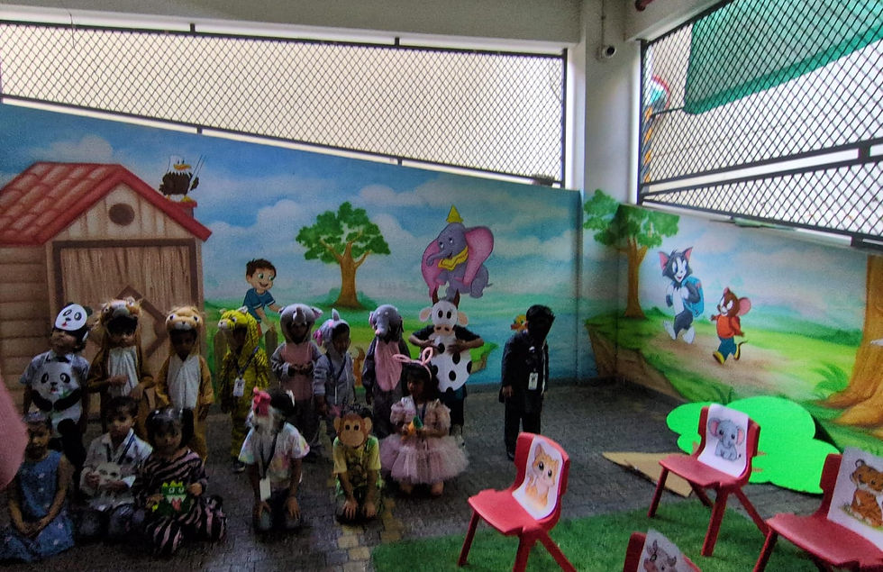 Children in animal costumes pose in a school courtyard with a cartoon mural and pink chairs labeled with animal pictures.