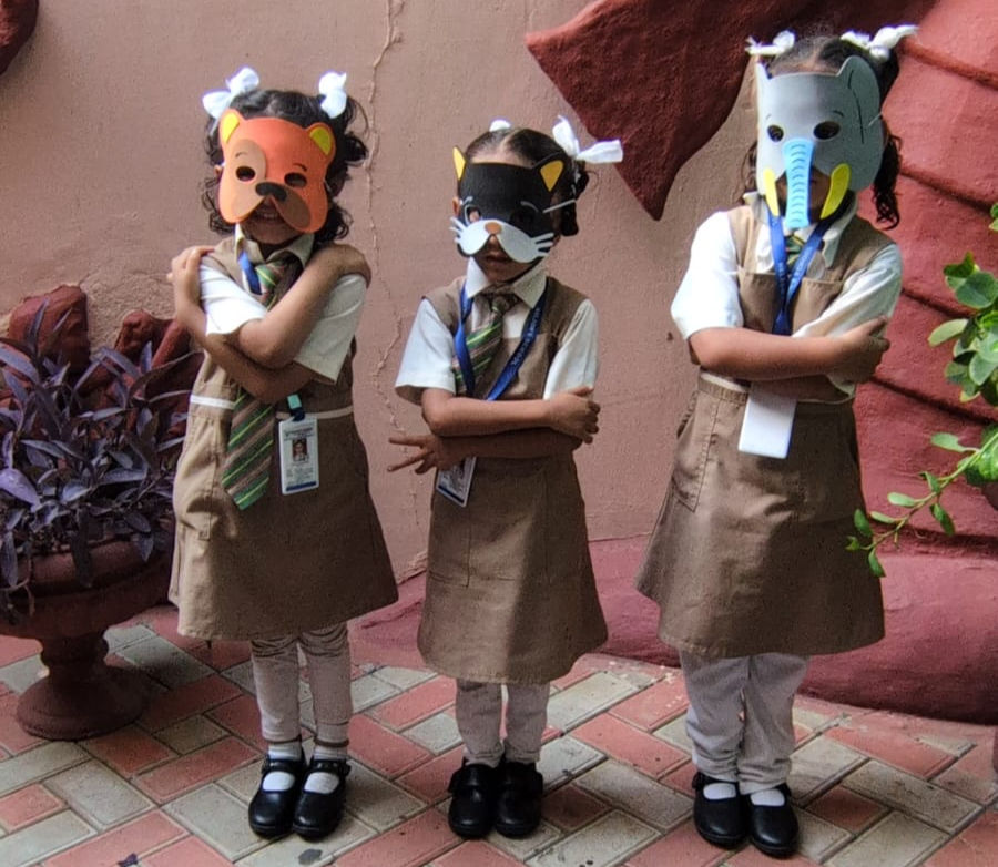Three schoolchildren in uniforms and animal masks stand with arms crossed in a pink-walled courtyard.