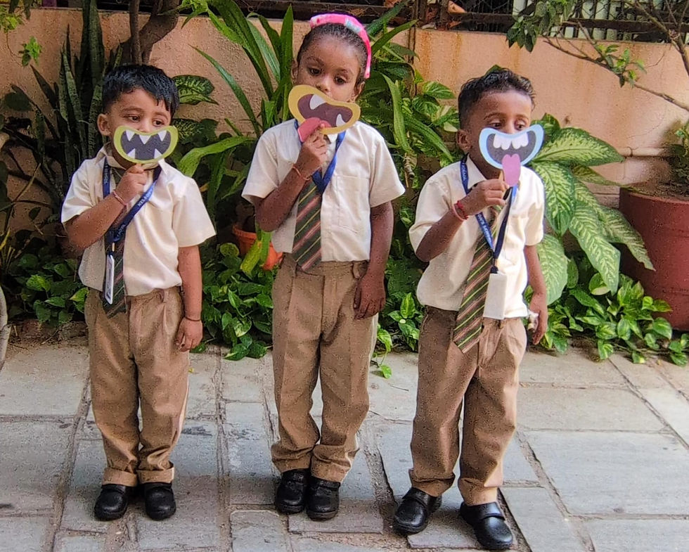 Three schoolchildren in uniforms hold colorful monster-face masks outdoors, standing on a paved path by lush green plants.