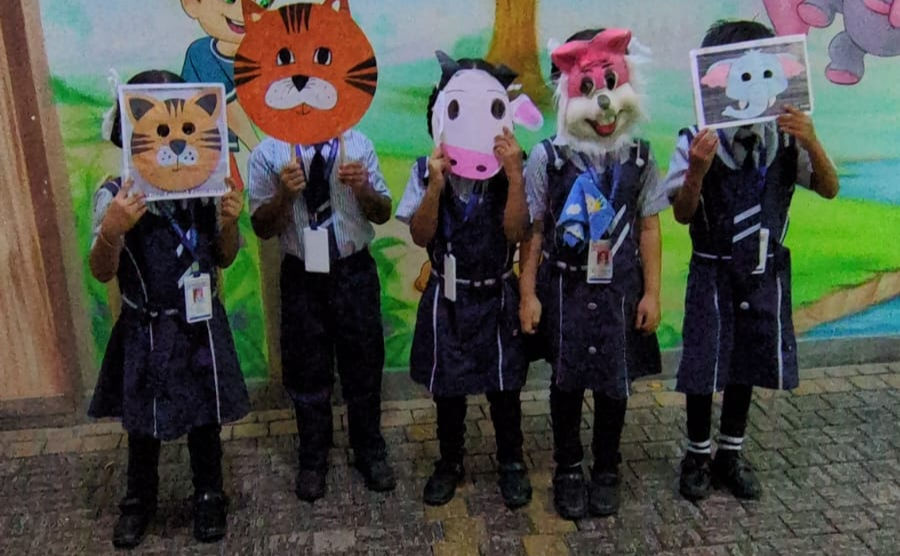 Five schoolchildren in uniforms hold animal face masks in front of a colorful mural, posing playfully outdoors.