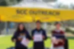 Three people smiling under a yellow "SCC Outreach" tent, holding papers on a sunny day, with trees and buildings in the background.