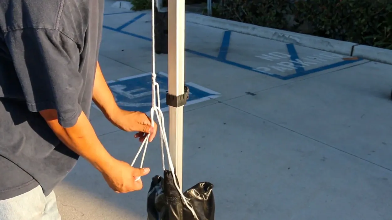 Hands looping a white rope around a canopy leg next to a black weight bag