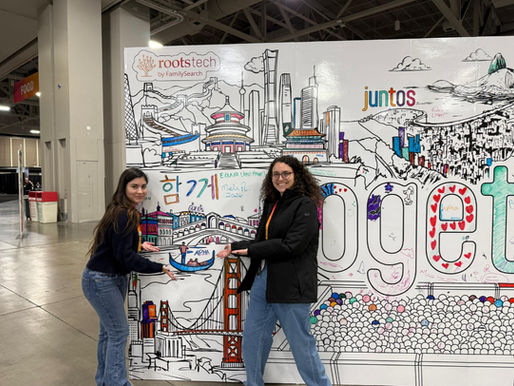 As genealogistas da Alpha Cidadania, Gabriela Rigazzo e Andrezza Calafate, posam sorridentes em frente a um grande mural interativo no RootsTech 2026. Elas apontam para um desenho de uma gôndola veneziana onde foi escrito o nome 'ALPHA'. O painel decorado apresenta ilustrações coloridas de marcos globais, como o Cristo Redentor e a Ponte Rialto, além das palavras 'juntos' e 'together' sob a marca oficial 'rootstech by FamilySearch', celebrando a presença da equipe no maior evento de genealogia do mundo.