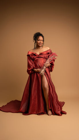 Elegant pregnant woman in red off-shoulder gown holding decorative leaves.