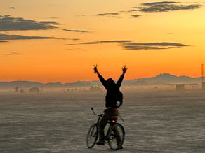 An attendee o a bicycle at Burning Man raises his arms to the sunrise in silhouette