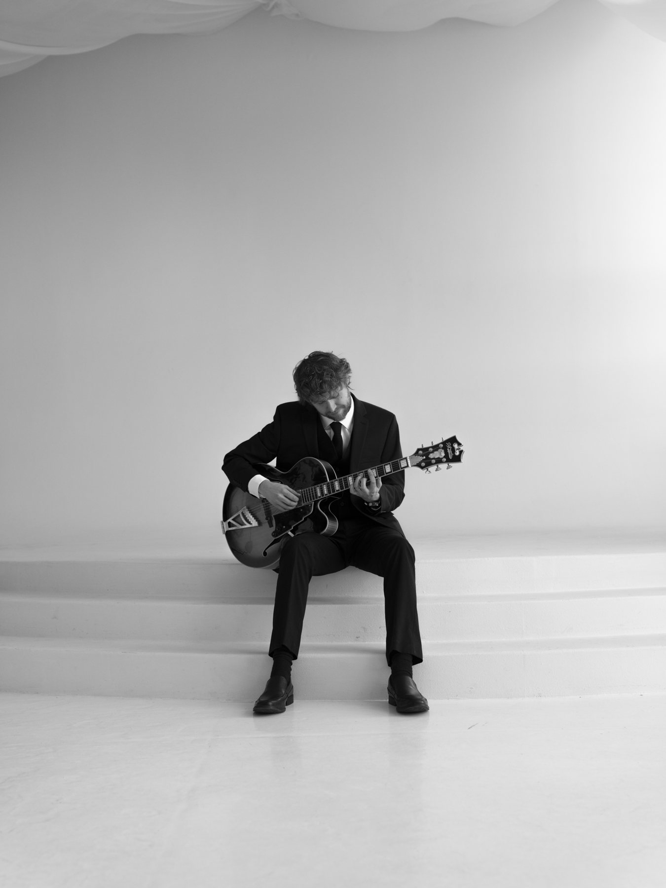 NYC jazz guitarist Hans Luchs in a dark suit playing an archtop guitar on white studio steps, black and white
