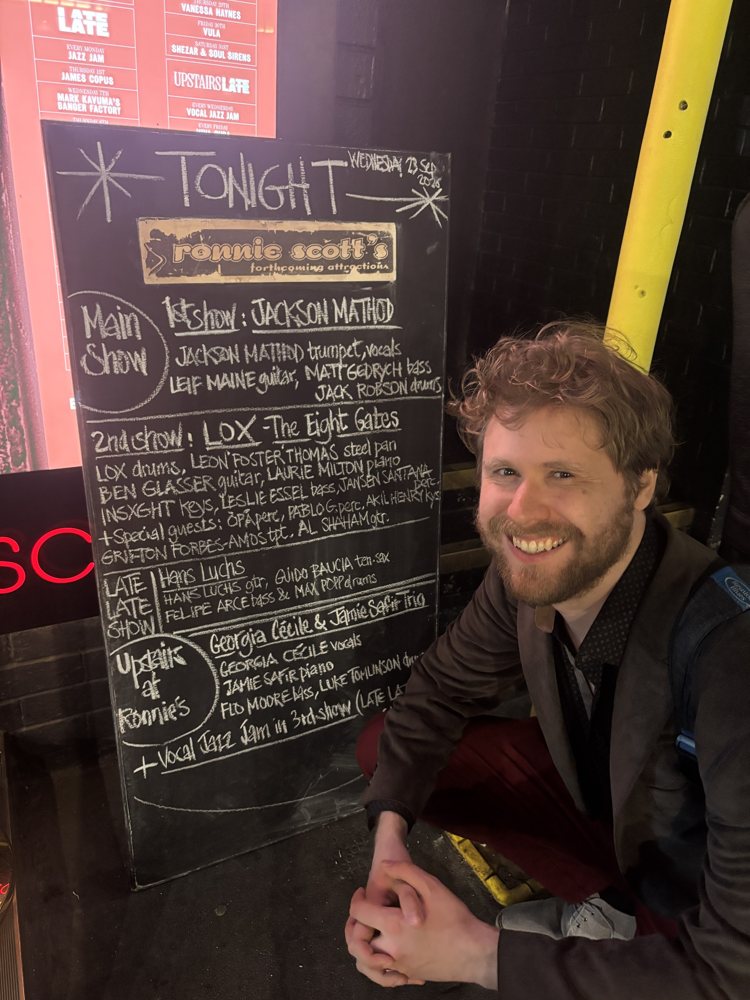Hans Luchs smiling beside the Ronnie Scott's chalkboard listing his Late Late Show