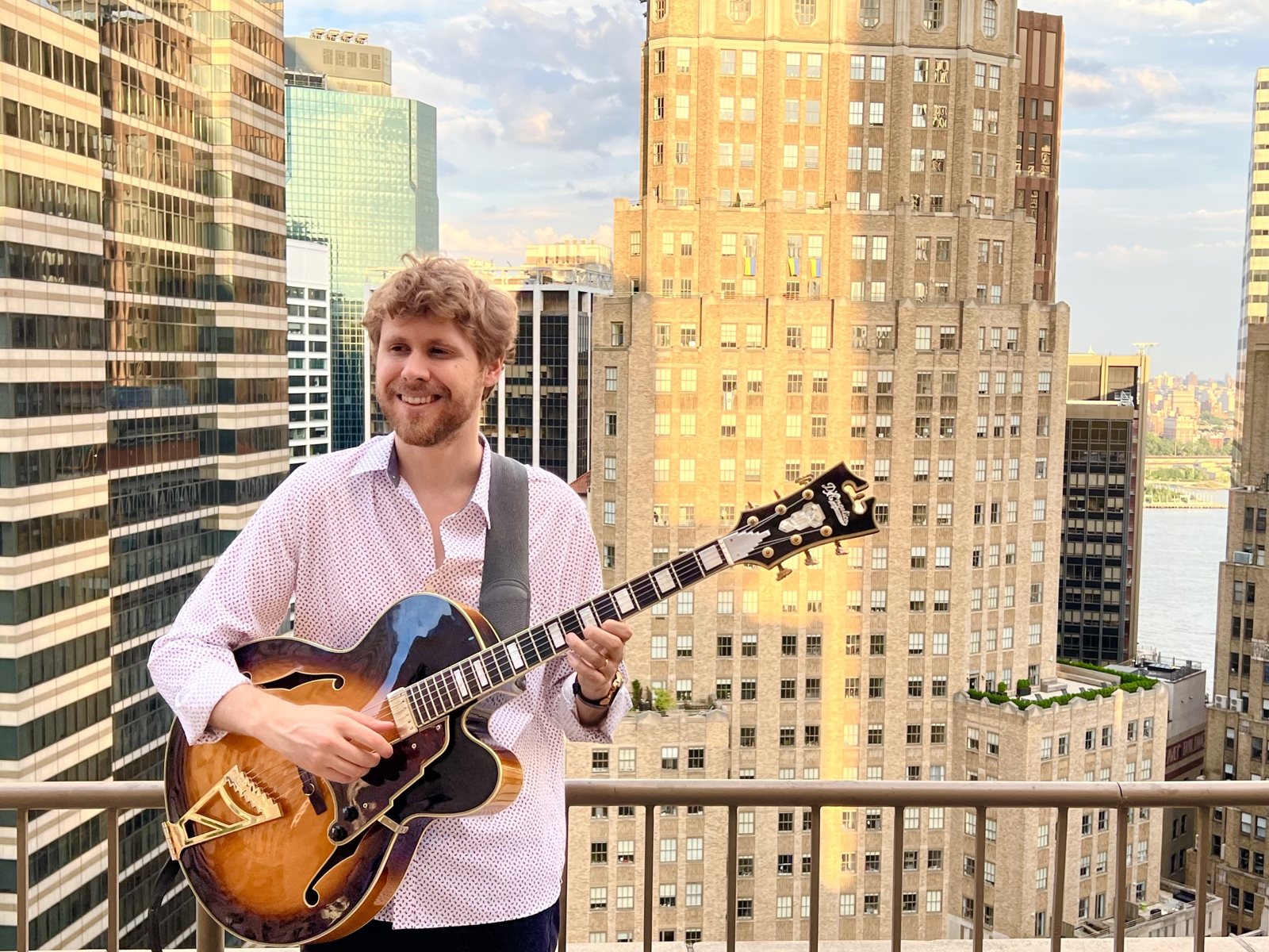 Hans Luchs smiling with his archtop guitar on a Manhattan rooftop at golden hour