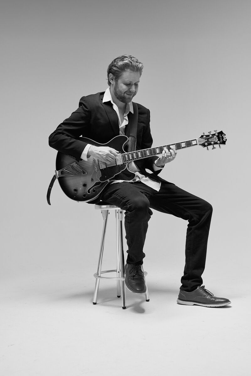 Hans Luchs seated on a stool with his archtop guitar, black and white
