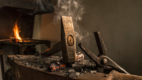 A worn blacksmith’s hammer resting upright on an anvil, surrounded by glowing embers, ash, and forging tools, with smoke rising in a dim workshop — a symbol of old-world craftsmanship and work forged by hand.