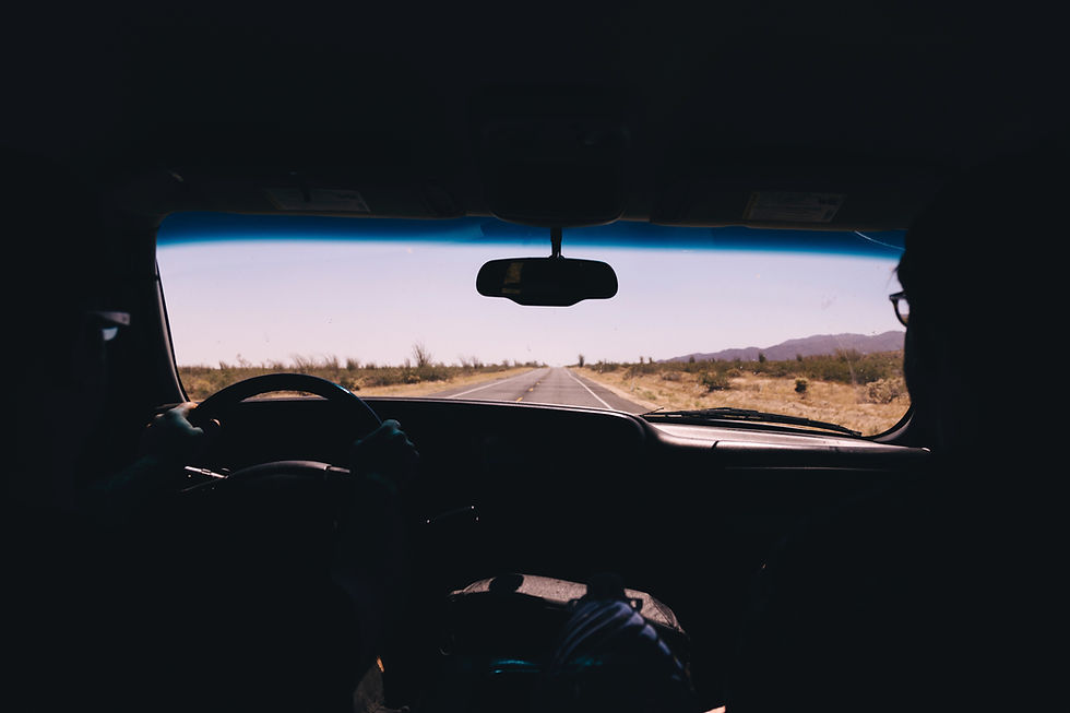 View from inside a vehicle traveling a quiet desert highway, with the driver and passenger visible in silhouette as the open road stretches toward the horizon.