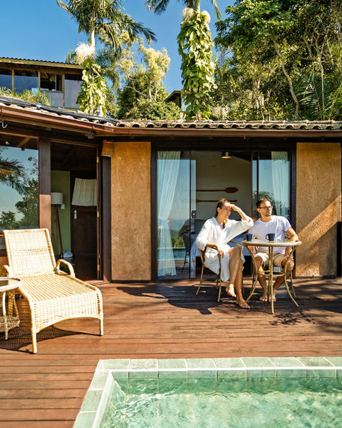 Casal relaxando na varanda de um chalé com deck de madeira e piscina privativa, cercado por vegetação tropical e céu azul em um ambiente tranquilo