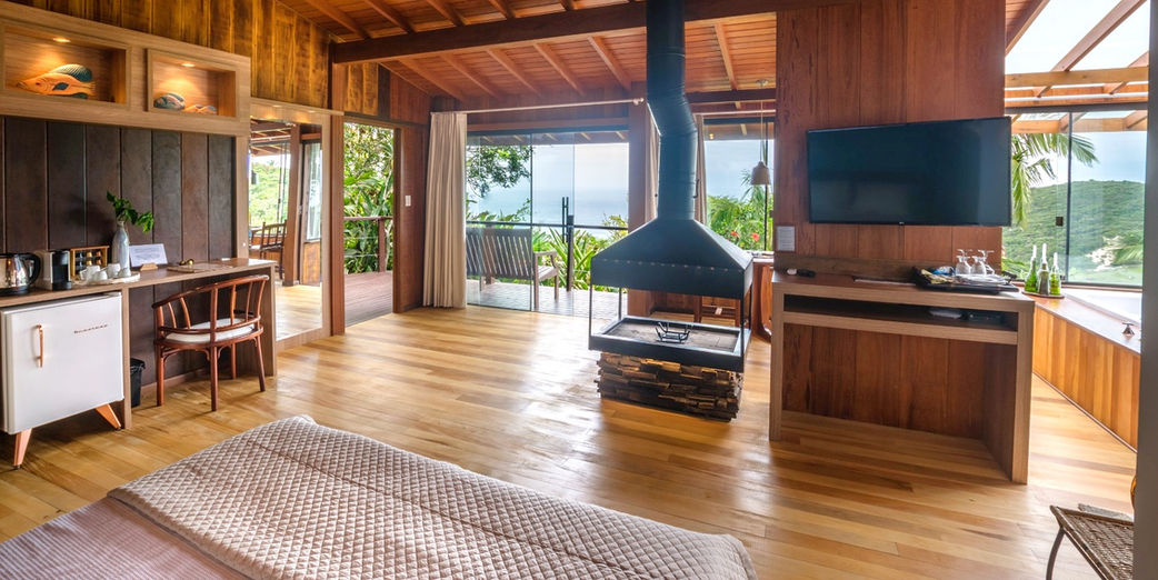 Visão Geral do Canto Esquerdo do Chalé Master Plus 1 com lareira de centro, SmartTV, mesa e frigobar retrô com bela vista do mar e montanha do Canto Sul da Praia do Rosa ao fundo