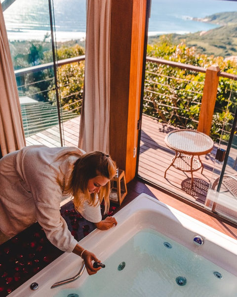 Mulher colocando sais e espuma de banho na banheira de hidromassagem do chalé com varanda e vista para o mar e natureza da praia do rosa