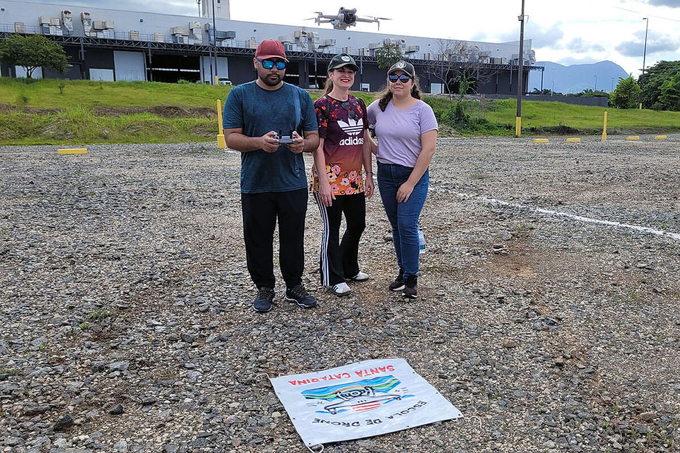 Três pessoas posam em terreno de cascalho, na Expoville, em Joinville SC, com um drone voando acima e uma faixa no chão com texto DRONE SANTA CATARINA.