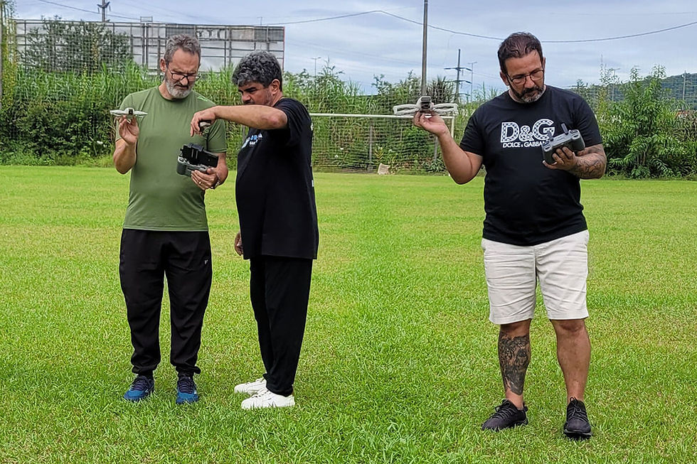 Três homens em campo gramado testam drones e controles, com foco concentrado; um veste camiseta D&G.