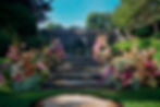 Colorful floral arrangements line cascading stone steps in a lush garden, surrounded by greenery and a bright blue sky.