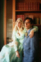 A couple poses in vintage attire; a woman in a mint dress and a man in a blue suit, in front of a bookshelf filled with leather-bound books.