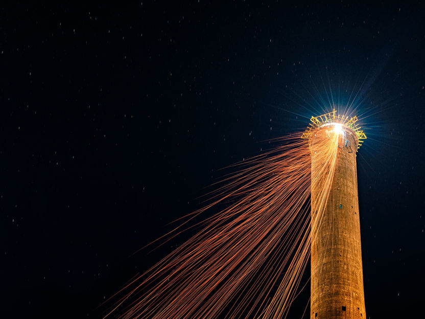 Chimney stack under construction at night with welding sparks creating artistic light trails beneath a star-filled sky.