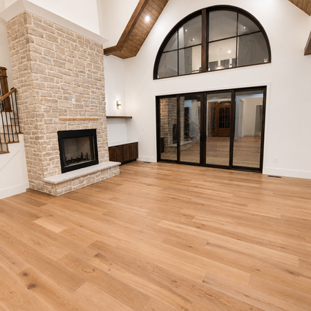 Grand vaulted family room featuring a floor-to-ceiling stone fireplace, exposed wood beam ceiling details, oversized arched window, expansive sliding glass doors, and wide-plank hardwood flooring in a bright open-concept layout.