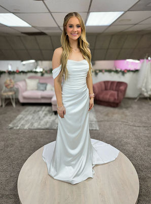 Bride wearing fit and flair wedding satin dress. With a swoop neckline embellished with pearls. This wedding dress ruches at the waistline to create a slimming look. It has off the shoulder straps to add to the romantic neckline. She is posing in a bridal shop. 
